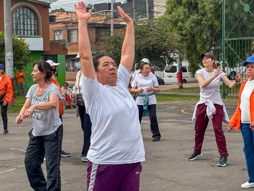 Grupo de personas adultas mayores realizando ejercicios al aire libre en una cancha del barrio. En el centro, una mujer con camiseta blanca levanta los brazos hacia arriba mientras otras personas a su alrededor estiran y mueven los brazos. Al fondo se observan edificios residenciales, árboles y una reja metálica.