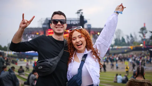 Fotografía de dos jovenes sonriendo y con las manos levantadas, de fondo desenfocado una de las tarimas del FEP y mucha gente alrededor