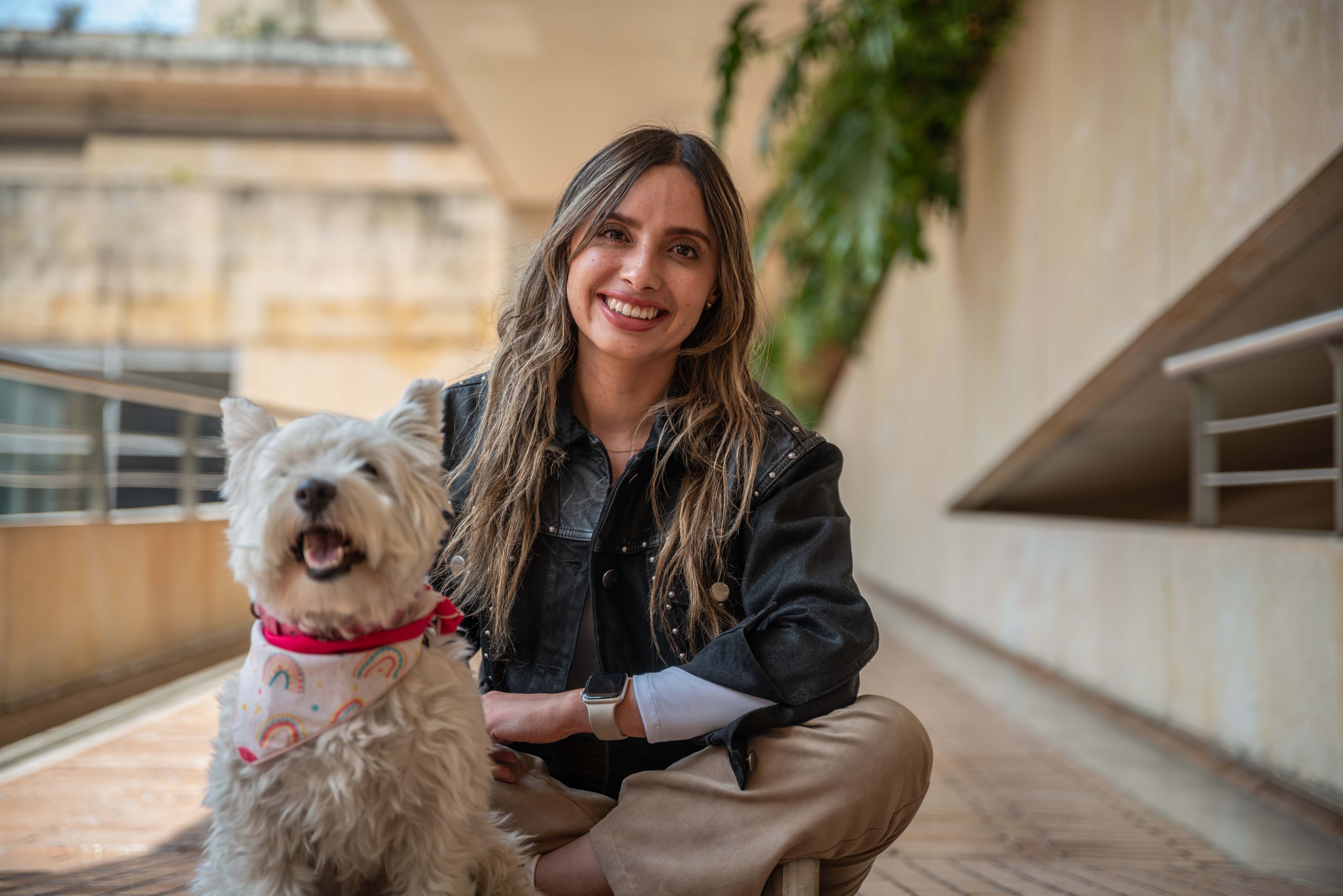 Foto de una mujer rubia jóven sentada y sonriendo, a su lado un perro blanco con collar rojo