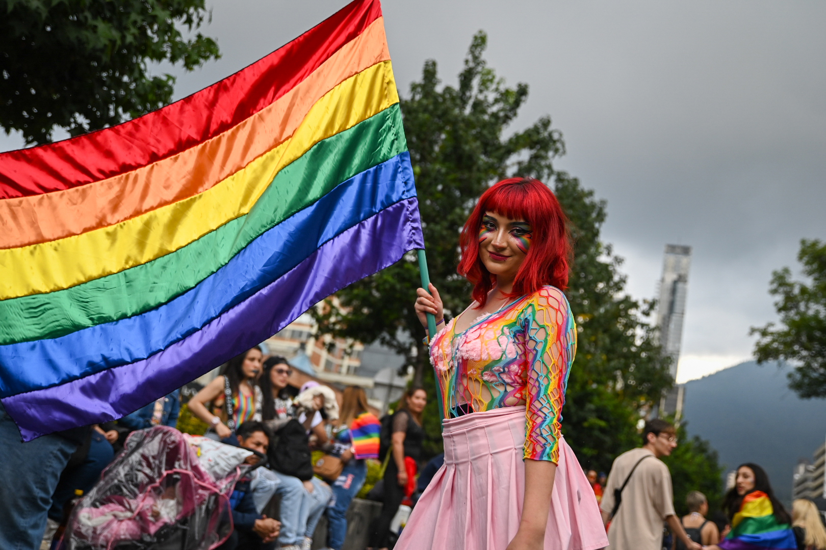 Foto de una mujer jóven sonrriente en su mano derecha sostiene la bandera de la comunidad LGTBI+