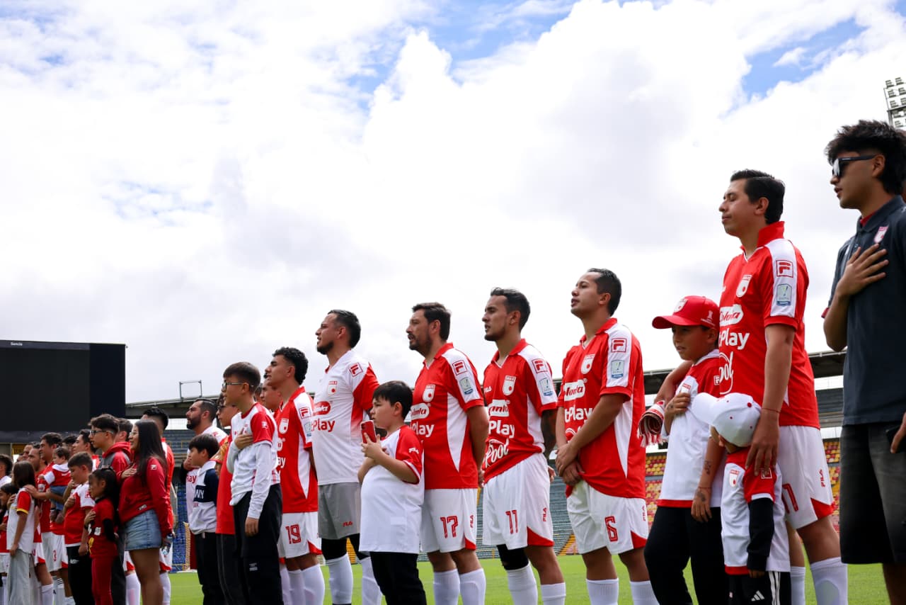 Foto de los jugadores de Santa Fe formados y en frente niñas y niños con camiseta de Santa Fe que los acompañan en la formación.