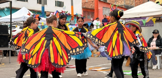 Grupo de niñas y jóvenes participa en una presentación cultural con coloridas alas de mariposa en un espacio comunitario al aire libre.
