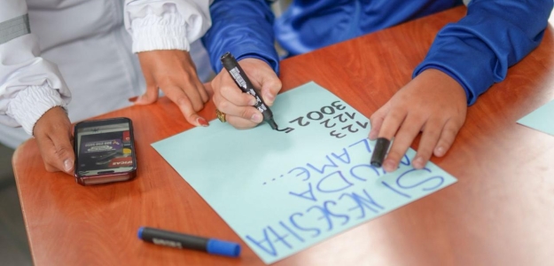 Foto donde se aprecian las manos de un menor de edad escribiendo nñumeros sobre una cartulina y a su lado se aprecian las manos de una persona adulta.