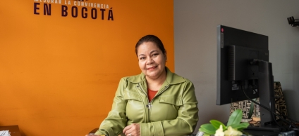 Inspectora de Policía en su escritorio sonriendo y en la pared del fondo de color mostaza se distingue el siguiente texto en blanco y negro "Trabajamos por mejorar la convivencia en Bogotá"