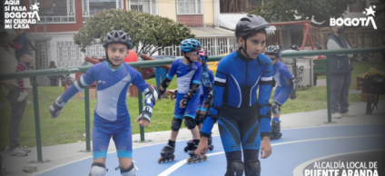 Niñas y niños practicando patinaje en una pista azul al aire libre mientras usan cascos y equipo de protección. Al fondo se observan viviendas, zonas verdes y personas acompañantes. La imagen muestra un ambiente deportivo y comunitario durante una jornada impulsada por la Alcaldía Local de Puente Aranda.