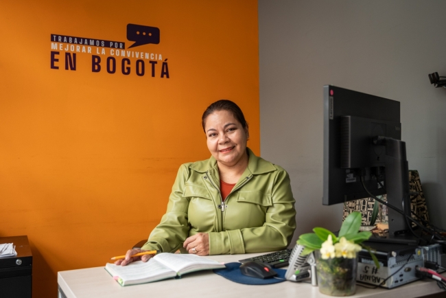 Inspectora de Policía en su escritorio sonriendo y en la pared del fondo de color mostaza se distingue el siguiente texto en blanco y negro "Trabajamos por mejorar la convivencia en Bogotá"