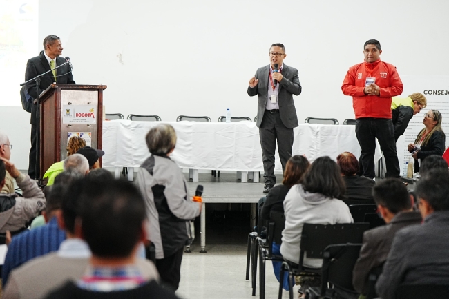 La imagen muestra un encuentro institucional con participación de la comunidad. En el escenario, varios representantes intervienen ante los asistentes durante una jornada de diálogo, mientras el público sigue la presentación desde el auditorio.