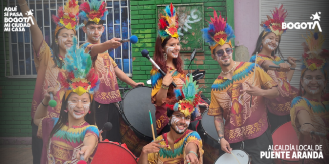 Grupo de jóvenes músicos posa sonriente en una calle, vestidos con trajes coloridos inspirados en culturas indígenas, con plumas y pintura facial. Sostienen tambores y baquetas, creando un ambiente festivo y cultural.
