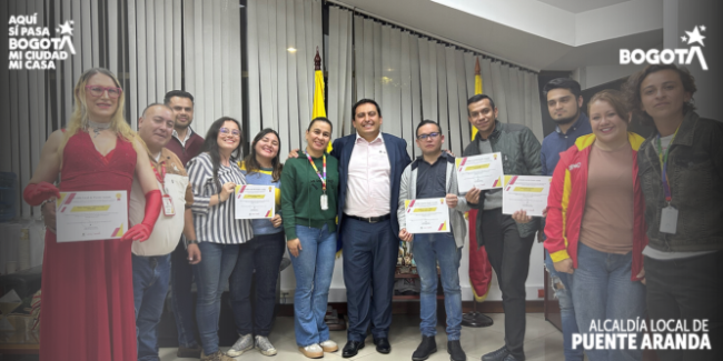 “Grupo de personas posando en un salón de la Alcaldía Local de Puente Aranda luego del acto de posesión del Consejo Local LGBTI 2026–2027. Varias de ellas sostienen certificados en las manos. En el centro se encuentra un funcionario de la Alcaldía acompañado por integrantes electos del Consejo. Al fondo se observan las banderas de Bogotá y de Colombia, y en las esquinas superiores aparecen los logos institucionales de ‘Bogotá’ y de la Alcaldía Local de Puente Aranda.”