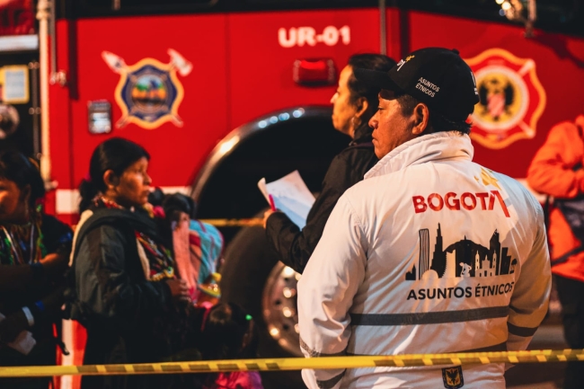 De fondo un carro de bomberos de Bogotá, se encuentra una mujer emebra haciendo fila y en frente dos funcionarios, uno de ellos de chaqueta blanca con el logo de Bogotá y el texto Diálogo
