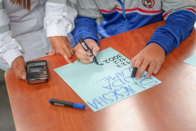 Foto donde se aprecian las manos de un menor de edad escribiendo nñumeros sobre una cartulina y a su lado se aprecian las manos de una persona adulta.