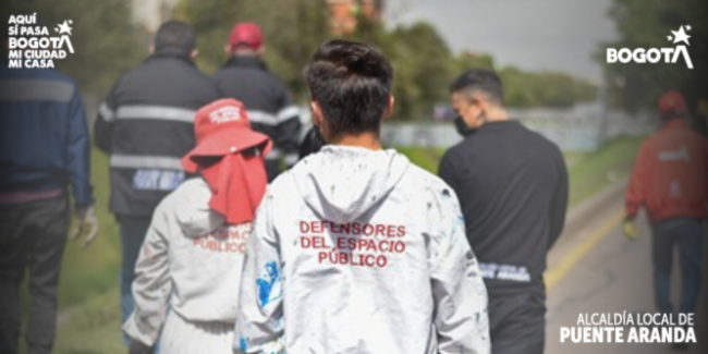 En la imagen se observan varios uniformados y gestores caminando por un corredor vial cercano a un canal, uno de ellos con chaqueta blanca que dice “Defensores del Espacio Público”.